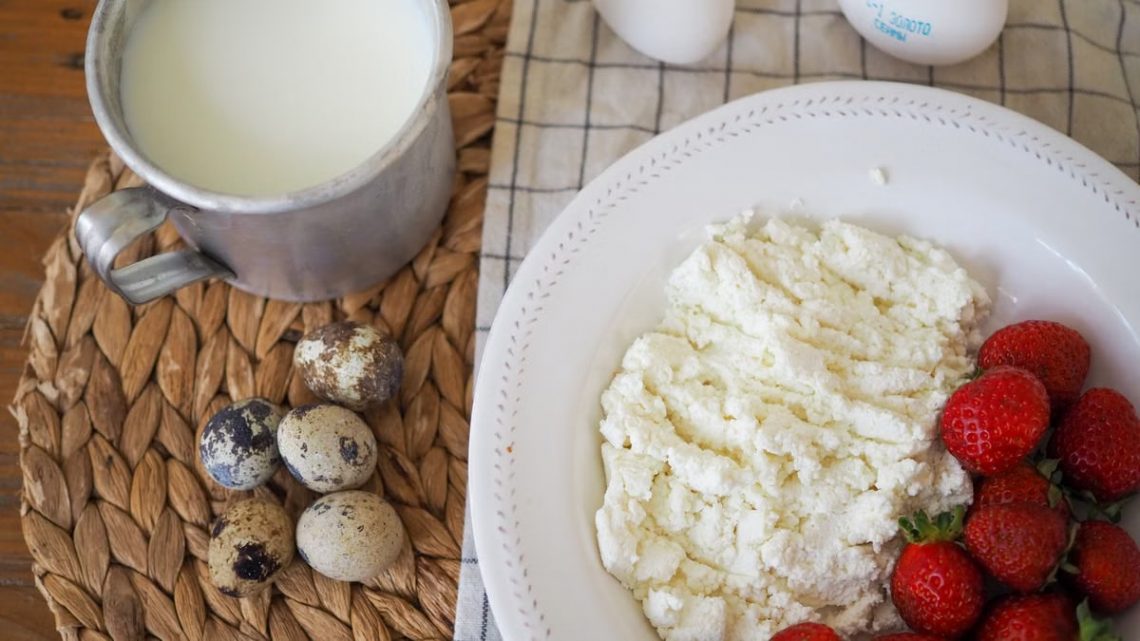 Top view of a rustic breakfast with milk, quail eggs, cottage cheese, and strawberries on a woven mat.